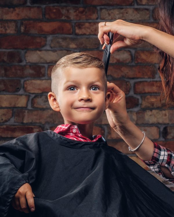 Smiling preschooler boy getting haircut. Children hairdresser with scissors and comb is cutting little boy in the room with loft interior.
